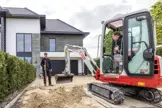 Hoveniers bezig met straat leggen, een zit in een minigraver, ander houdt schep vast